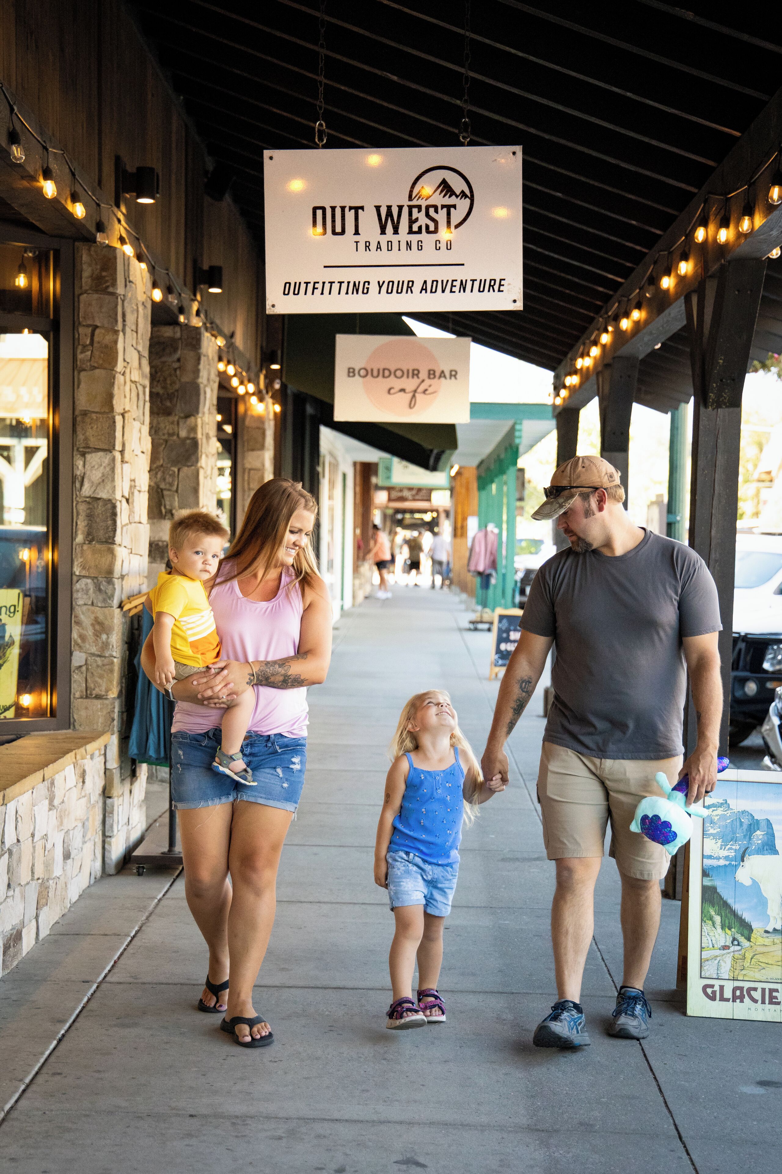 A family walking along a covered outdoor area with a sign for 'OUT WEST TRADING CO' and a 'BOUDOIR BAR'.