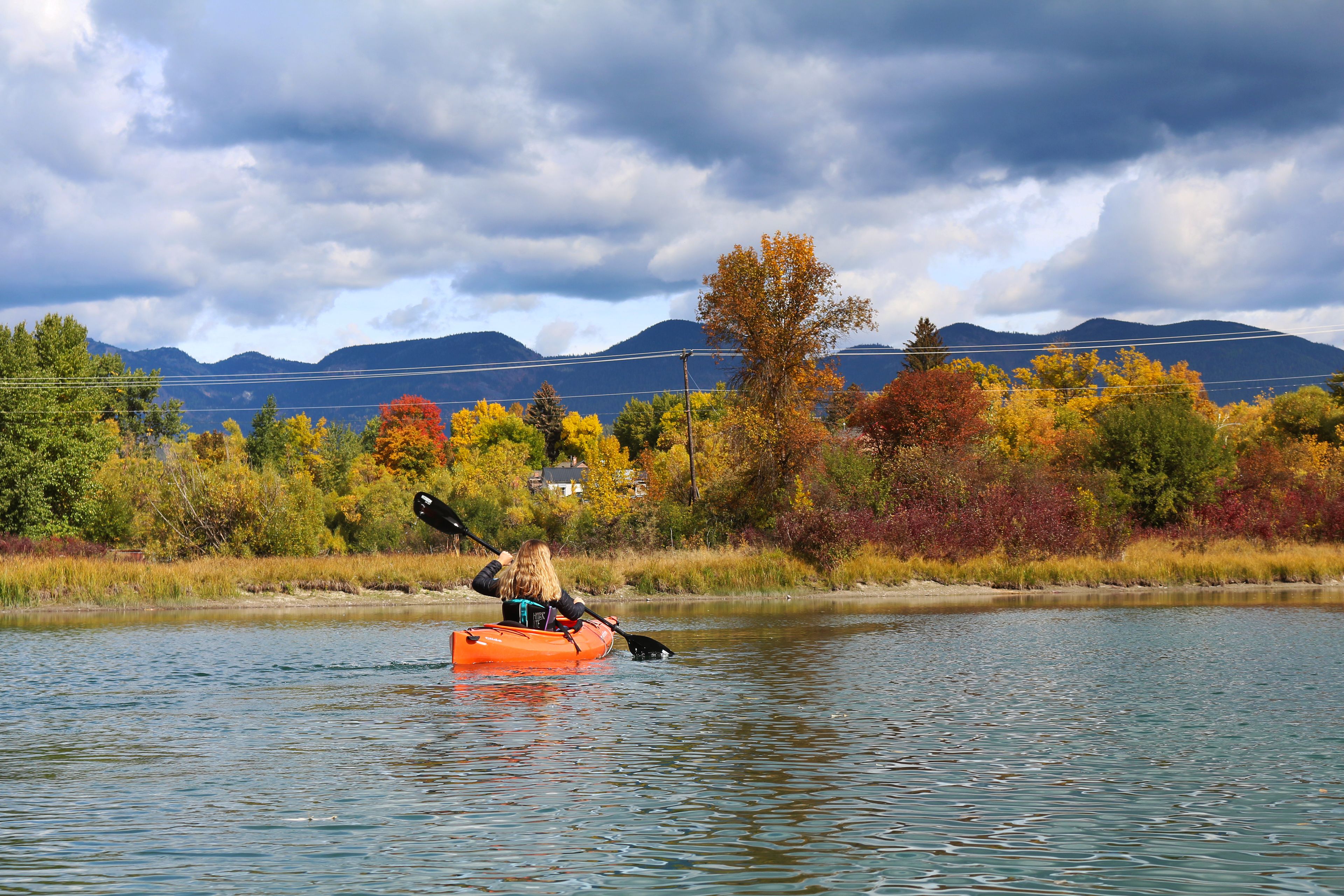 Una persona en kayak en un río, con follaje otoñal y montañas al fondo.
