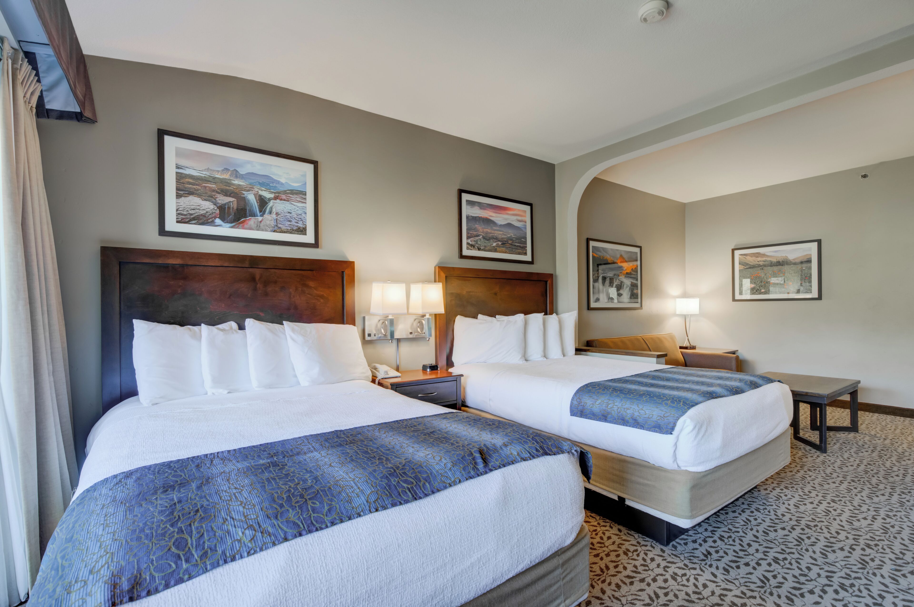 Une chambre d’hôtel avec deux lits, un bureau et une chaise. Les murs sont ornés de tableaux encadrés et le sol est recouvert d’une moquette à motifs.