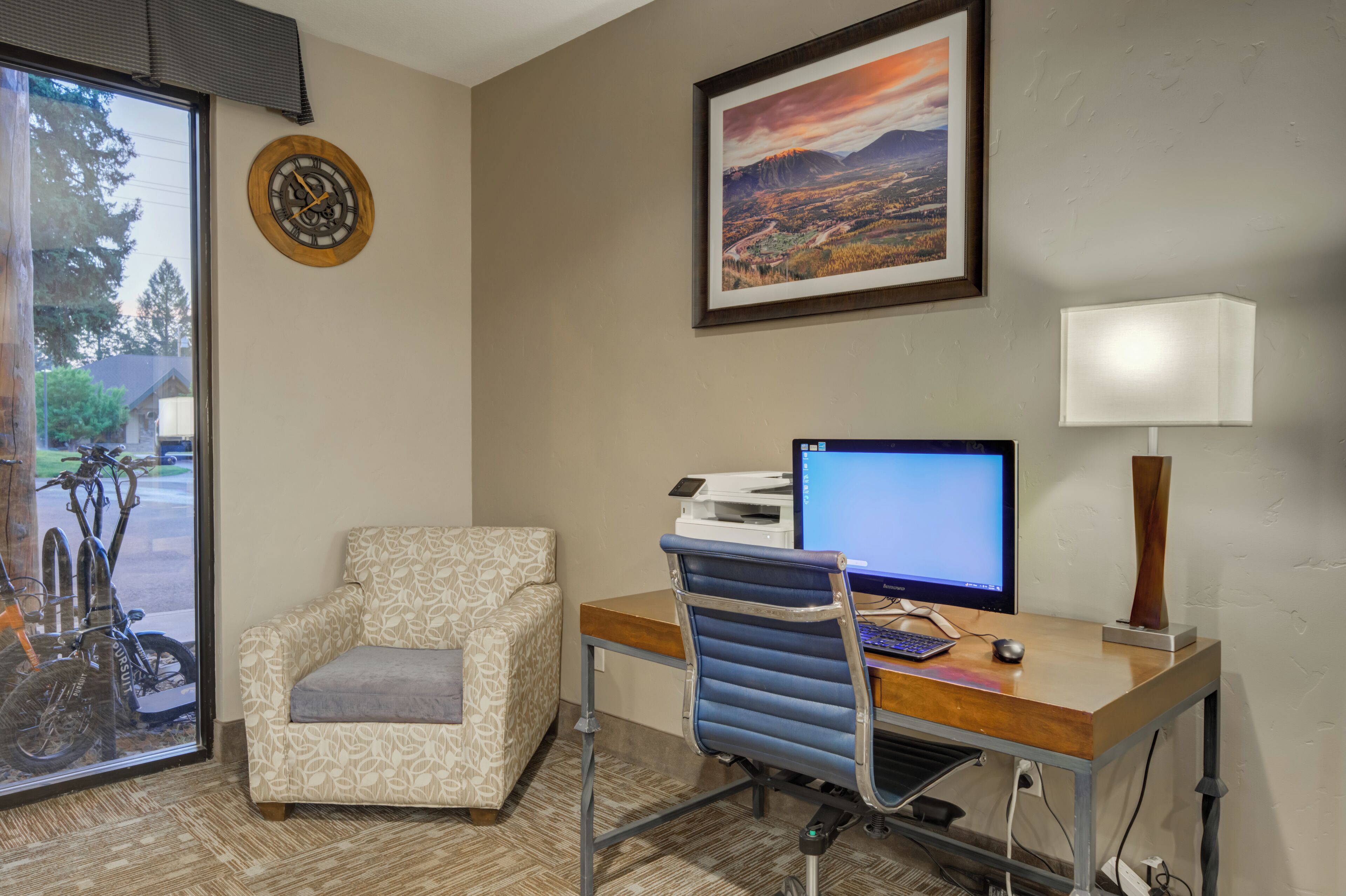 A room with a desk, computer, chair, lamp, clock, and a bicycle outside the window.