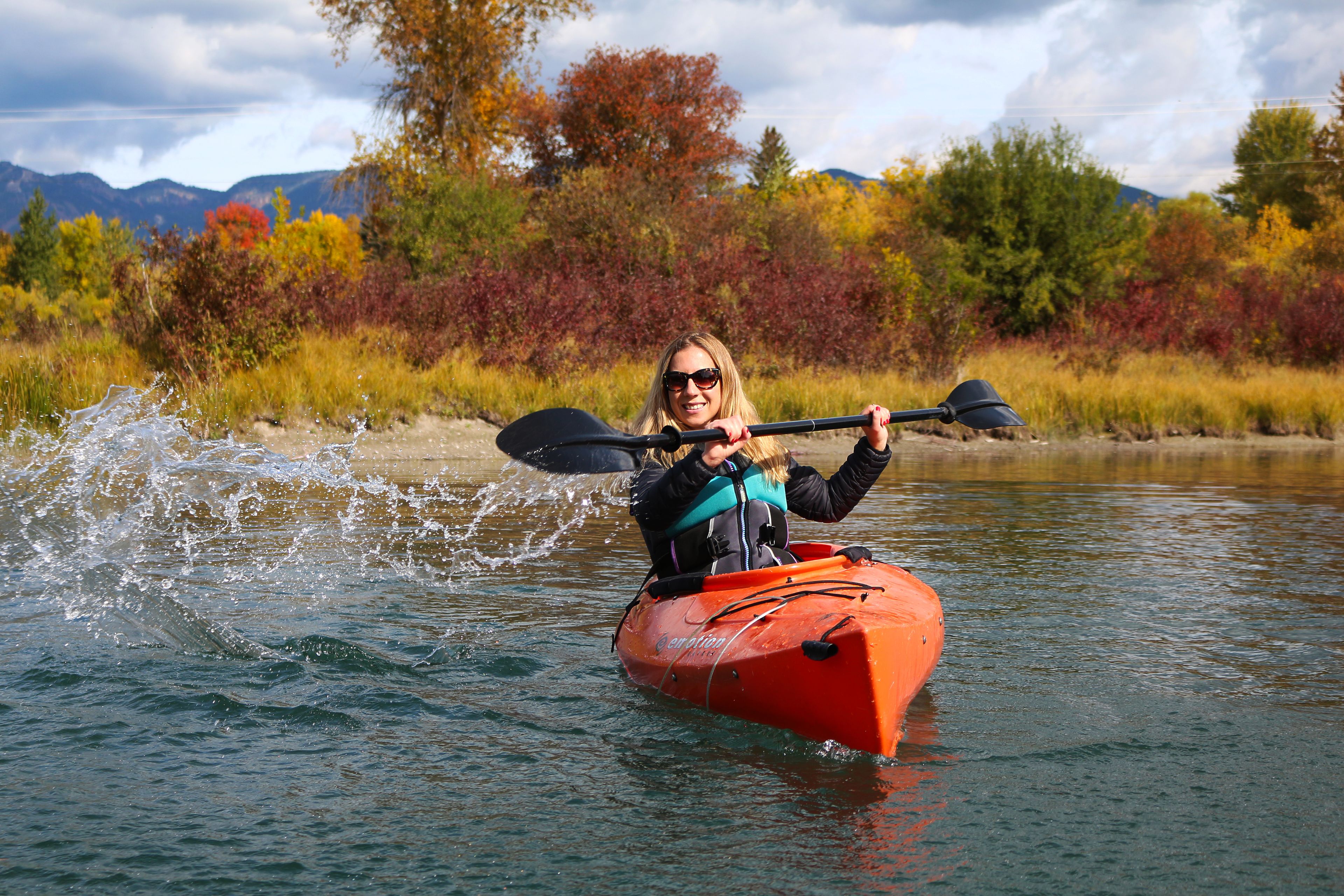 A person in an orange kayak on a river with autumn foliage in the background.