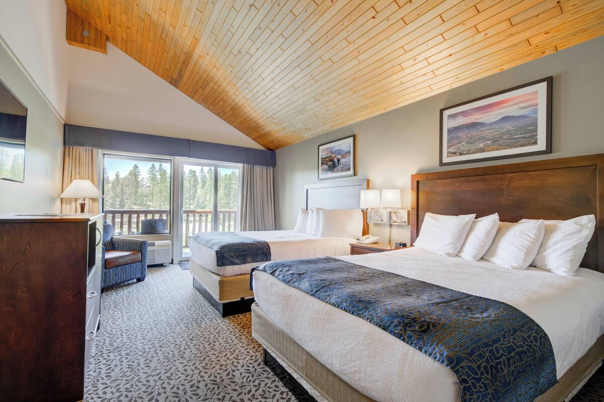 Een hotelkamer met twee bedden, een houten plafond, een groot raam met uitzicht op bomen, en een aan de muur hangend schilderij.