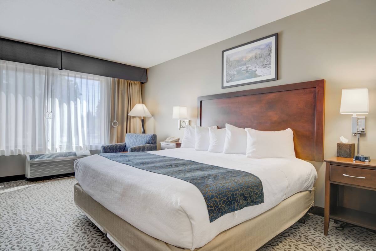 Een hotelkamer met een groot bed, twee nachtkastjes met lampen, een raam met gordijnen, een telefoon aan de muur en een lijst met een afbeelding boven het bed.