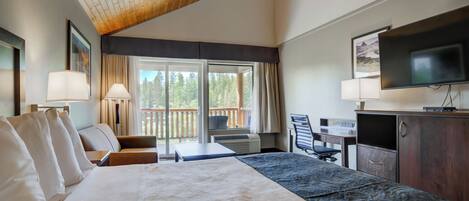Habitación de hotel con una cama grande, un escritorio, una silla, un televisor y un balcón con vistas a árboles.