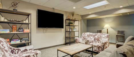 Un salón con televisor de pantalla plana, una mesa de centro y dos sofás con diseño de vaca. Hay estanterías con libros y objetos decorativos.
