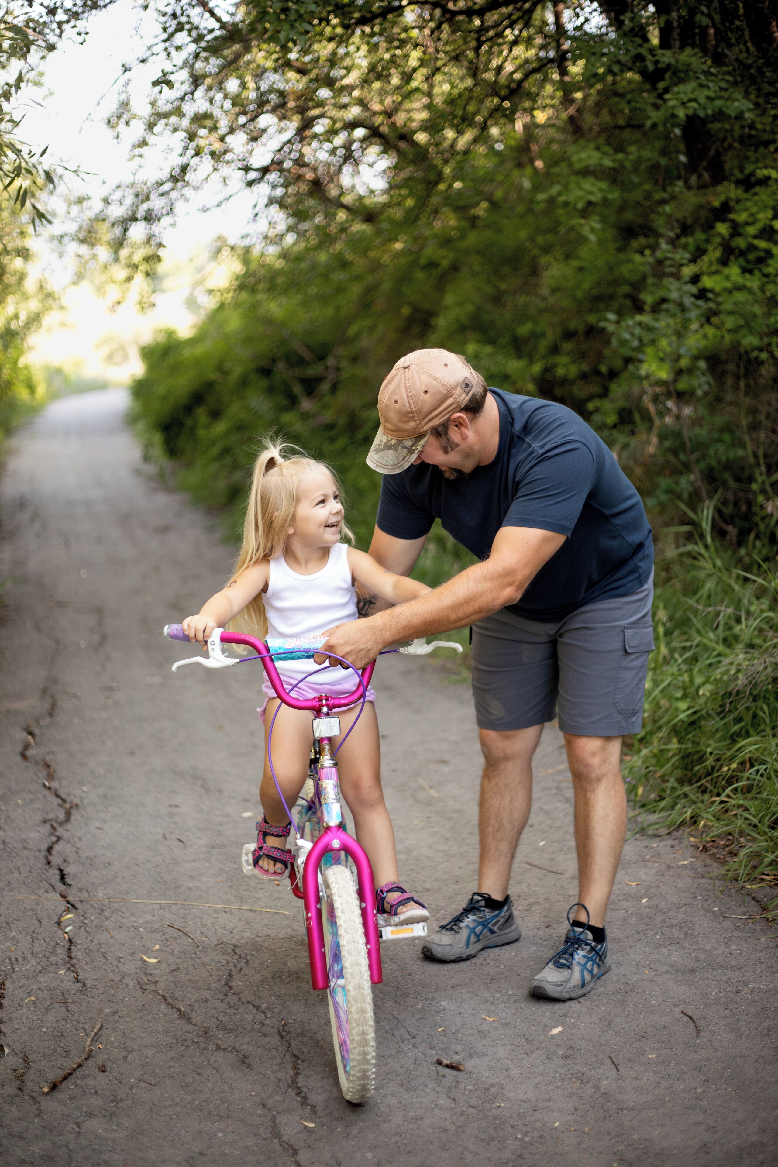 A man assisting a child on a pink bicycle on a paved path surrounded by greenery.