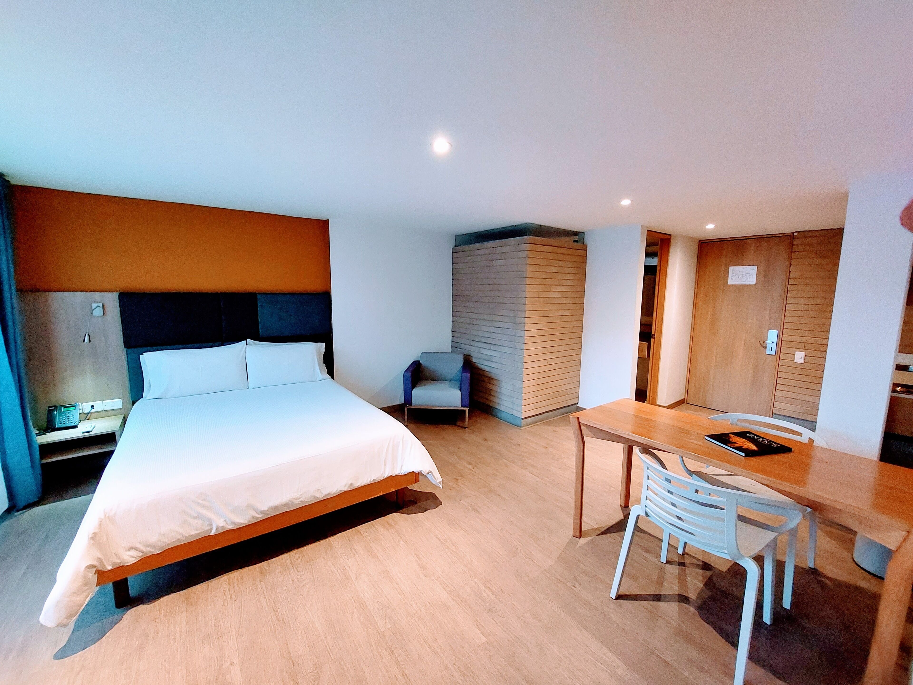 A modern hotel room with a large bed, a desk, and a chair. There is a small table with a book on it, and a wall-mounted phone.