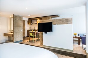 A modern hotel room with a large bed, a flat-screen TV mounted on the wall, a dining area with a table and chairs, and a kitchen area with a counter and appliances. - Viaggio Teleport Suites (Bogotá)
