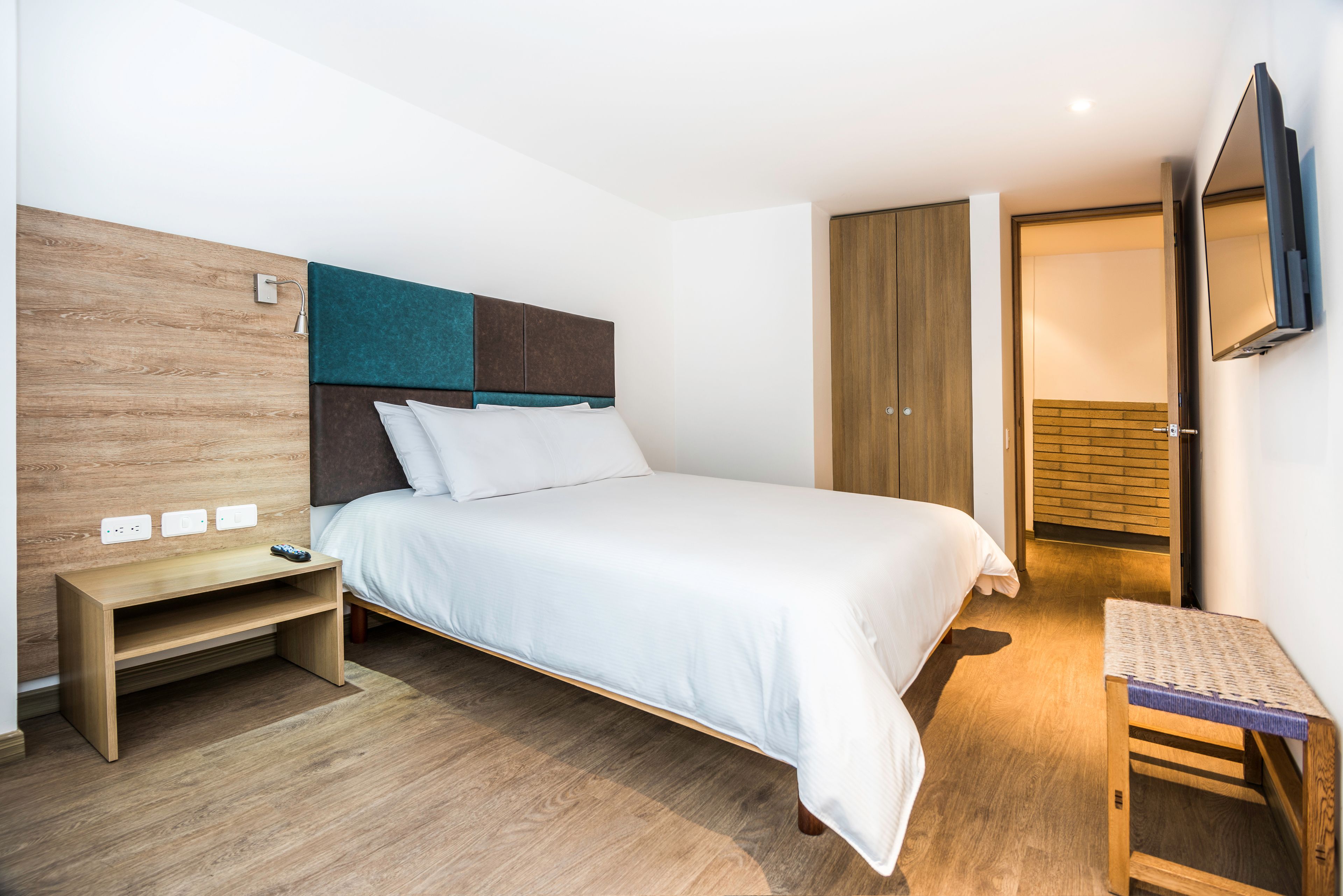 Ein modernes Hotelzimmer mit einem Bett, einem Nachttisch aus Holz, einem kleinen Tisch, einem Kleiderschrank und einem an der Wand befestigten Fernseher.