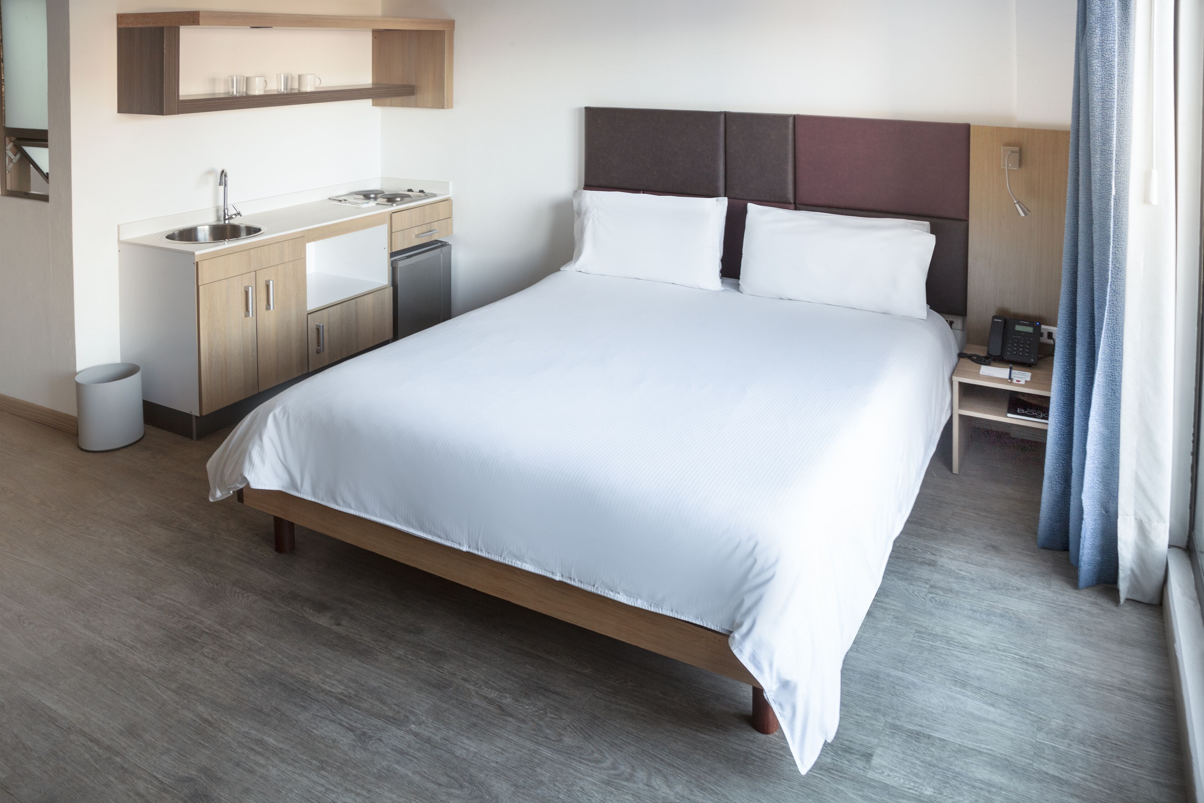 Una habitación de hotel moderna con una cama grande, cabecera de madera, una pequeña kitchenette con un lavamanos y una mesita de noche con un teléfono y una lámpara.