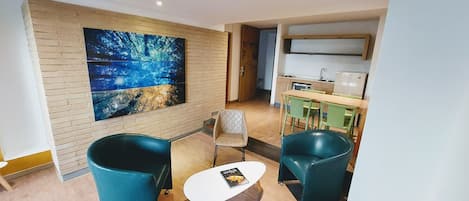 A modern living room with a large painting, two teal armchairs, a small table, and a kitchen area in the background.