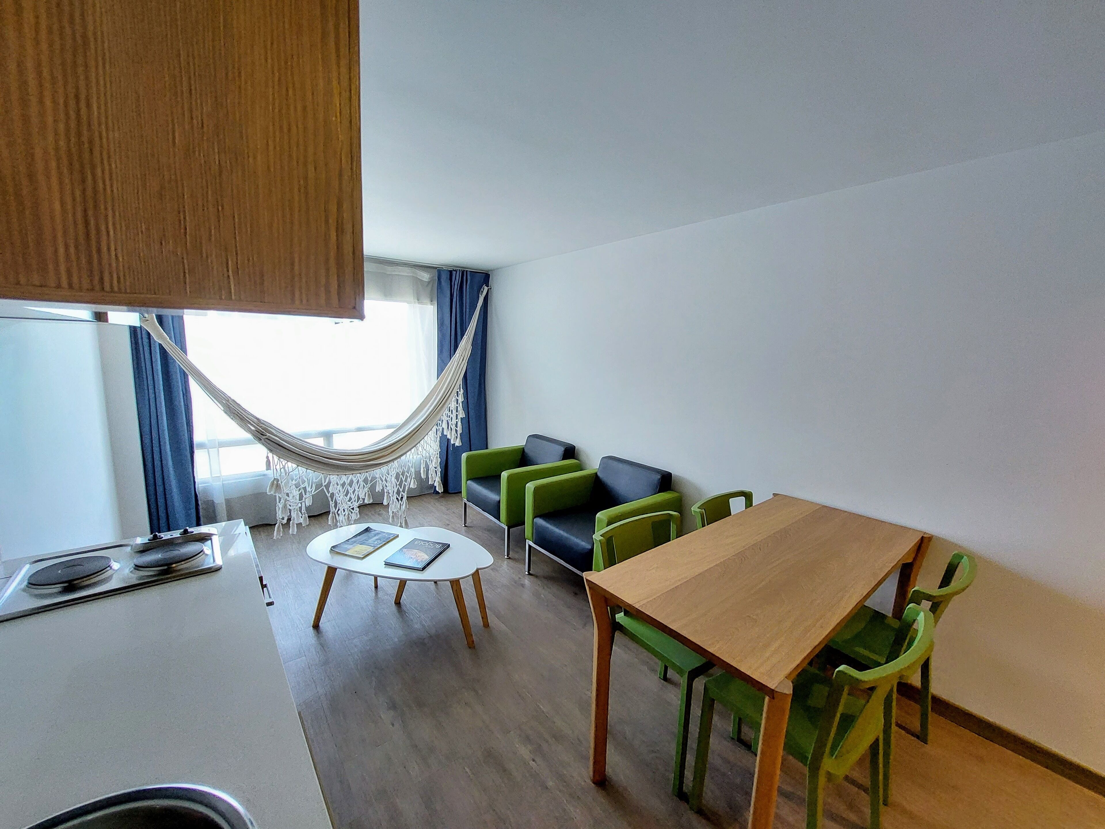 Una cocina moderna con una mesa y sillas de madera, una pequeña mesa redonda con libros y una hamaca junto a la ventana.