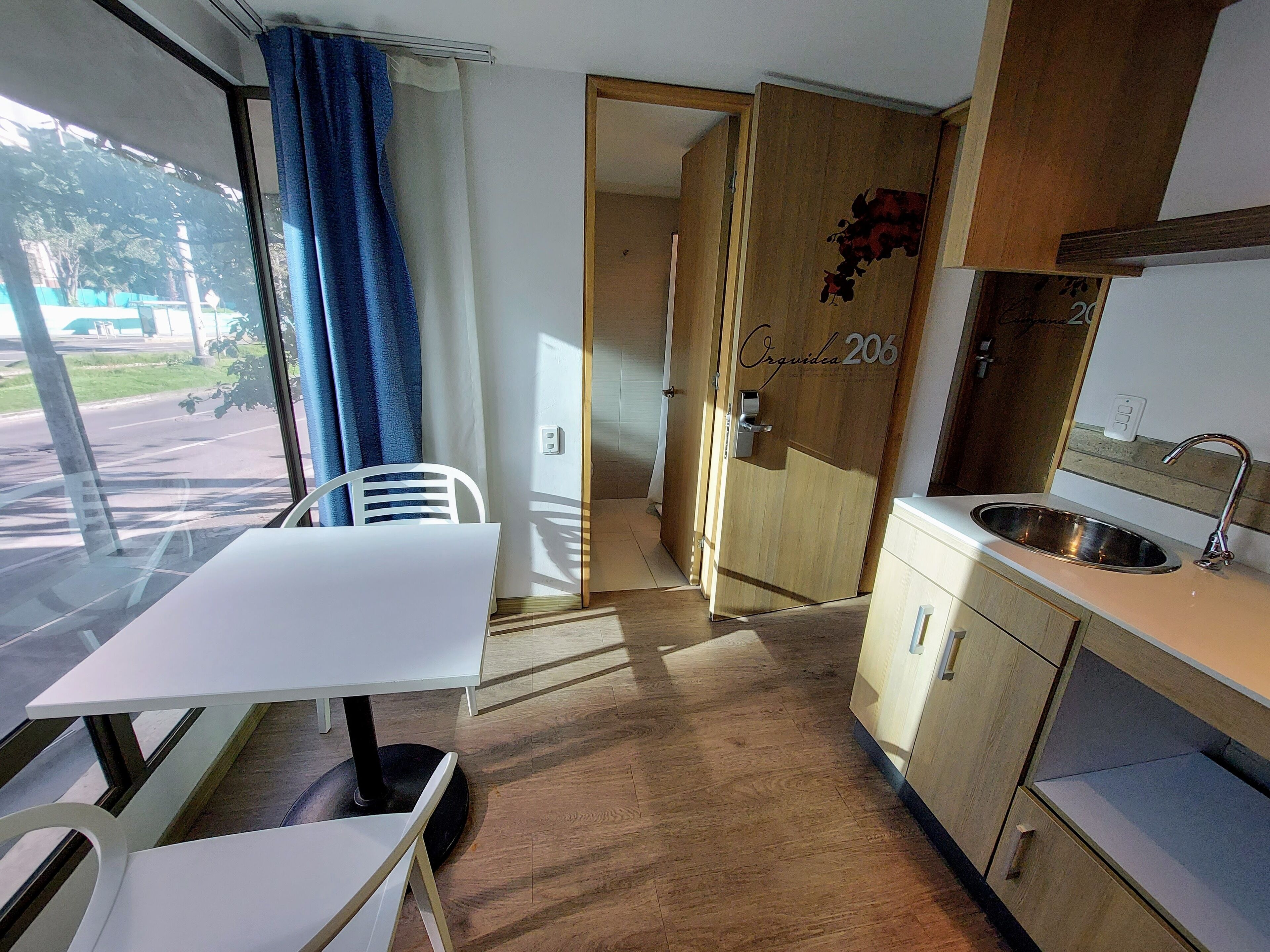 Una habitación con una mesa de comedor, una silla blanca, una puerta de madera con la etiqueta "Orquidea 206", una zona de cocina con un lavamanos y vista a la calle.