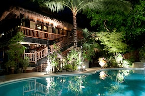 A poolside area with a thatched-roof structure, a wooden staircase, and lush greenery.