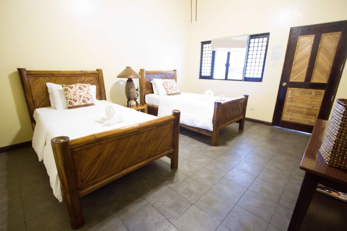 Une chambre d’hôtel avec deux lits en bois, une table de chevet, une lampe et une porte ornée d’un motif tissé.