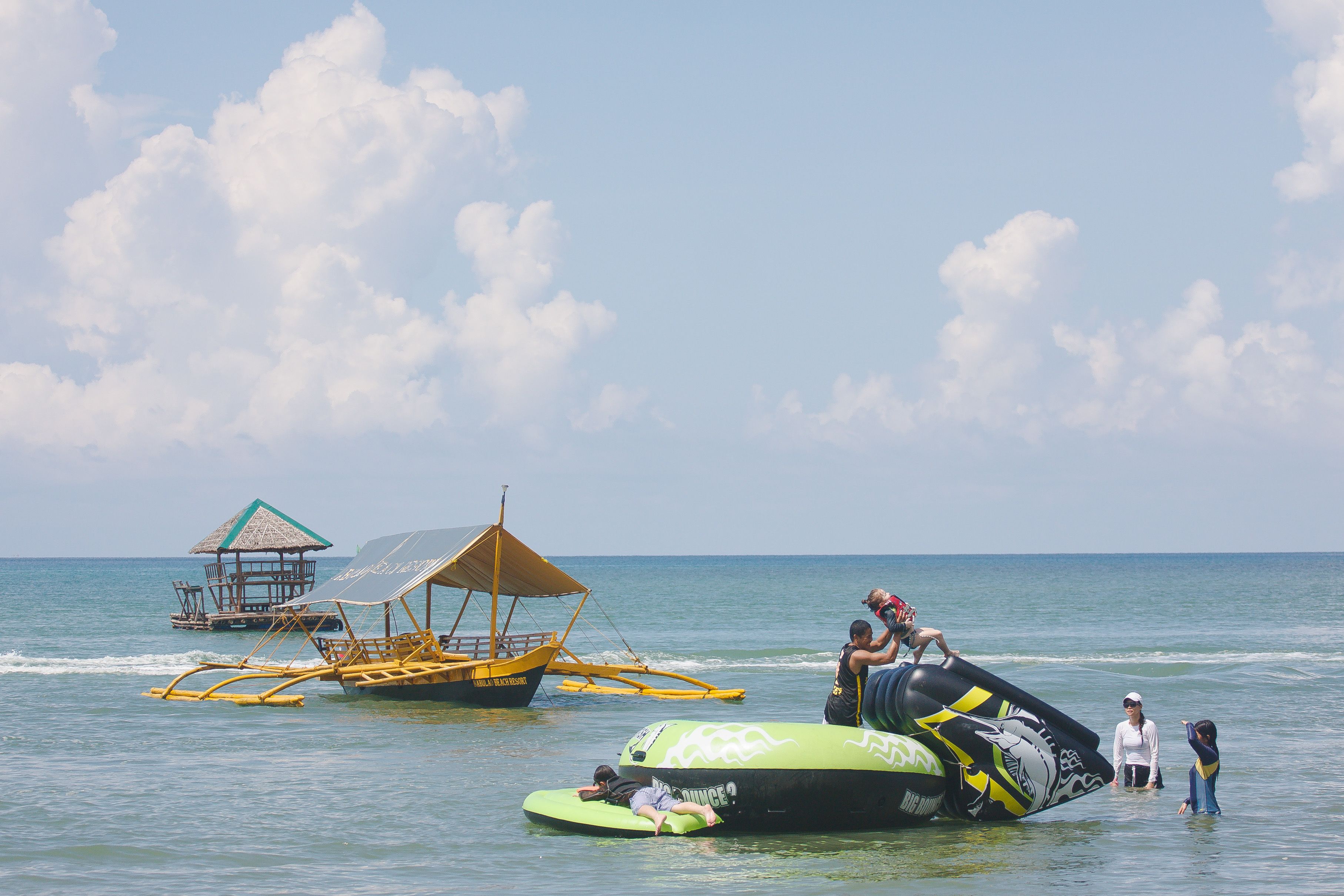 Une scène de plage avec un bateau en bois, une structure à toit de chaume et un radeau gonflable vert sur lequel se trouvent des personnes.