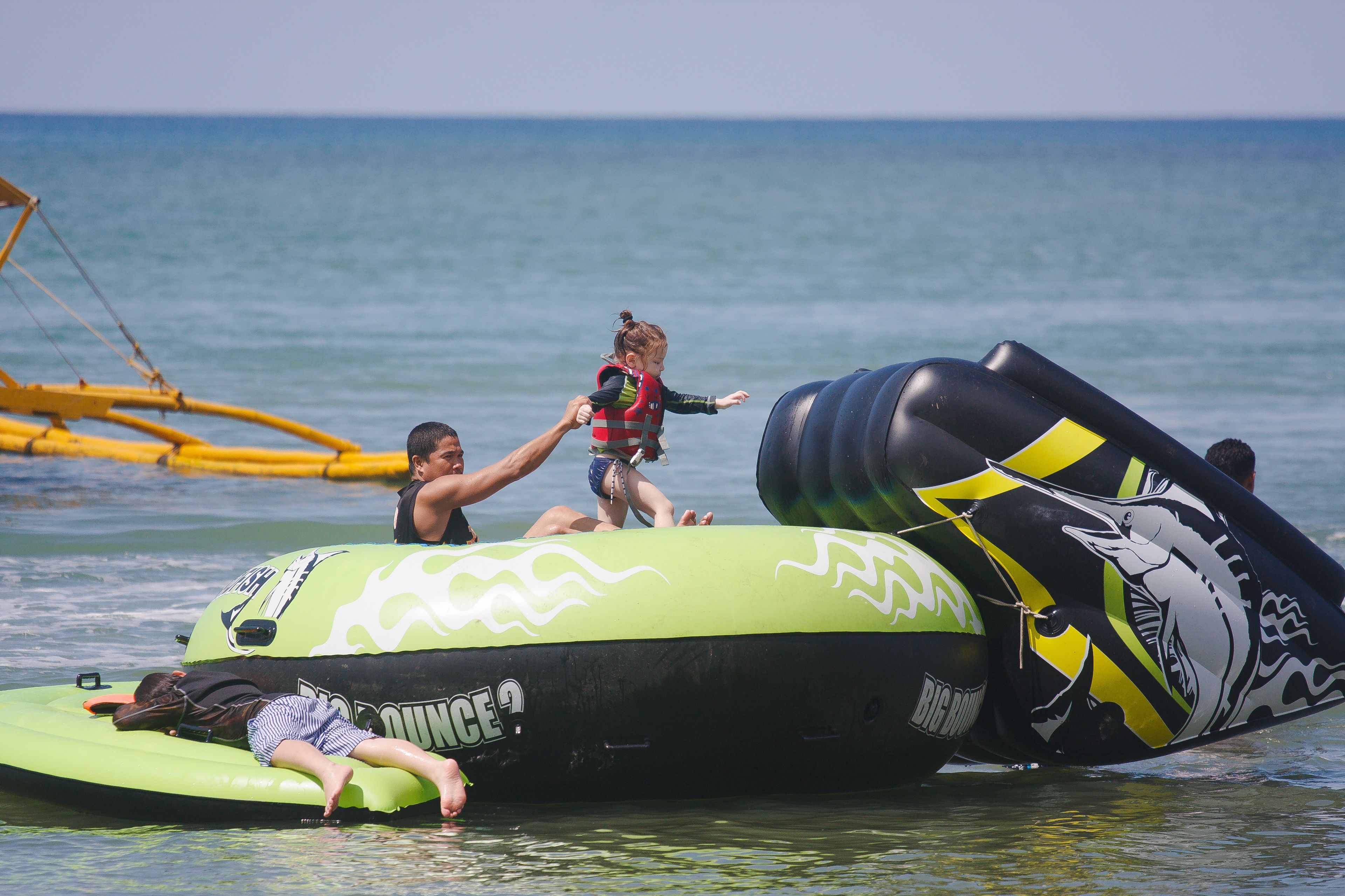 A person lying on a green inflatable raft in the water, another person standing on it, and a child in a life jacket.