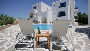 A poolside area with two lounge chairs, a small table with a bottle and glasses, and a white building with blue accents in the background. - Sunny Beach Studios (Naxos)