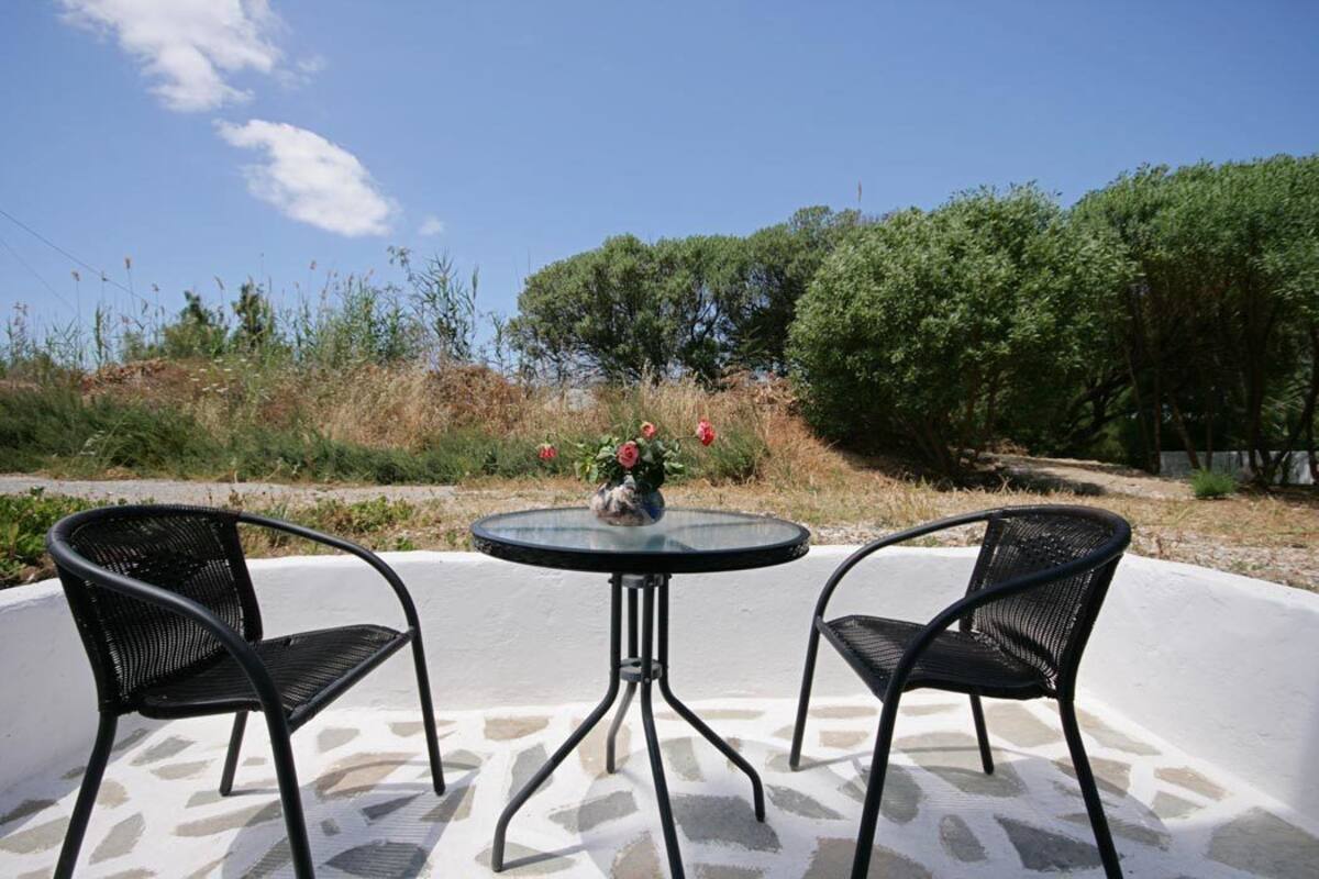 A patio with two black metal chairs and a glass table, surrounded by a white wall and a garden with trees and plants.