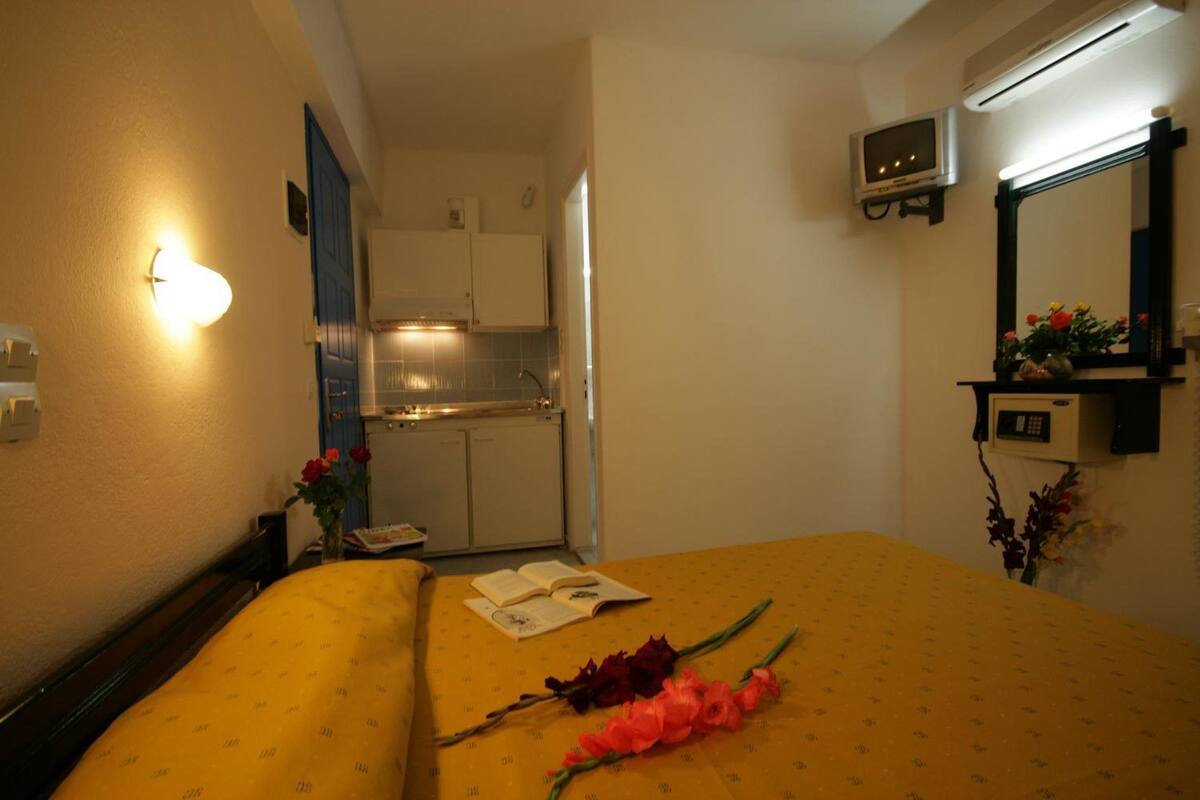 A hotel room with a bed, a small desk, a mirror, a TV, and a kitchenette in the background.