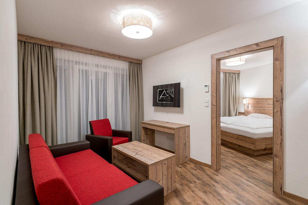 A hotel room with a wooden floor, a bed, a desk, a chair, and a TV.