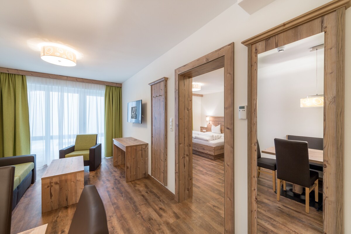 A hotel room with a wooden floor, a dining area with a table and chairs, a sofa, a bench, and a bed with a nightstand.
