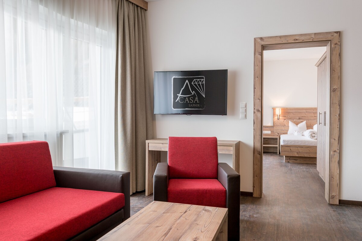 A hotel room with a TV mounted on the wall, a wooden desk, a red and black chair, a sofa, and a wooden floor.