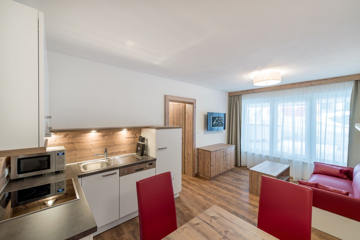 A compact kitchen with modern appliances, a dining area with red chairs, and a living space with a sofa and curtains.