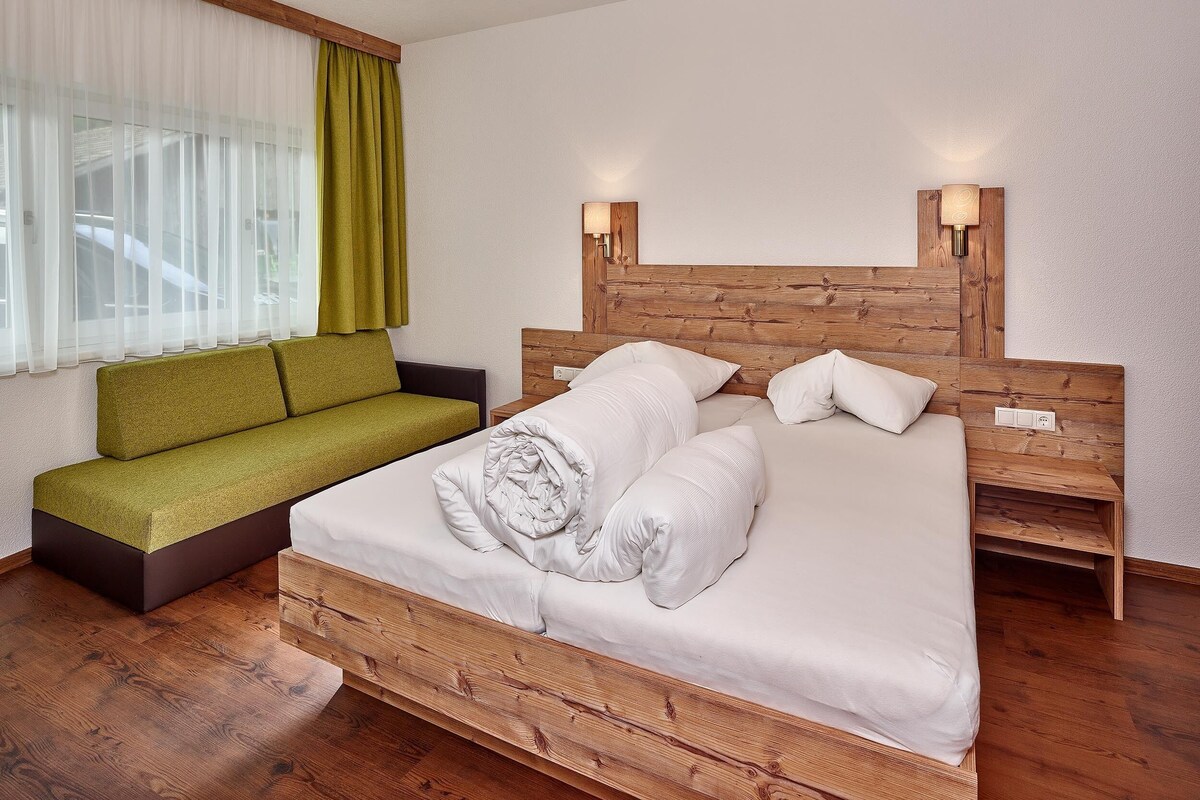 A bedroom with a wooden bed, a green sofa, a window with sheer curtains, and a wall-mounted light fixture.