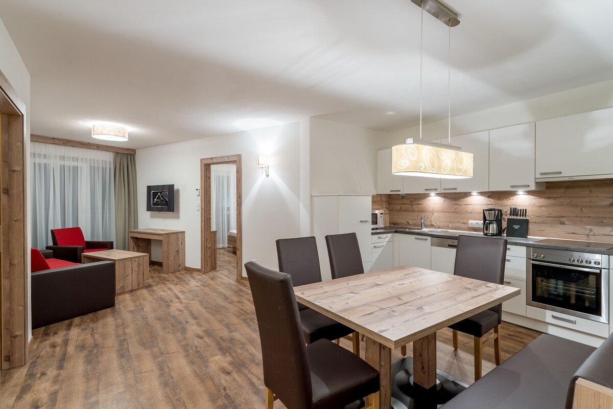 A modern kitchen with white cabinets, a wooden countertop, and a built-in oven. There is a dining area with a wooden table and chairs, and a small seating area with a red chair.