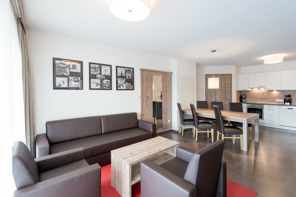 A modern living room with a sofa, a coffee table, and a dining area with chairs and a table. There is a kitchen area with appliances and a hanging light fixture.