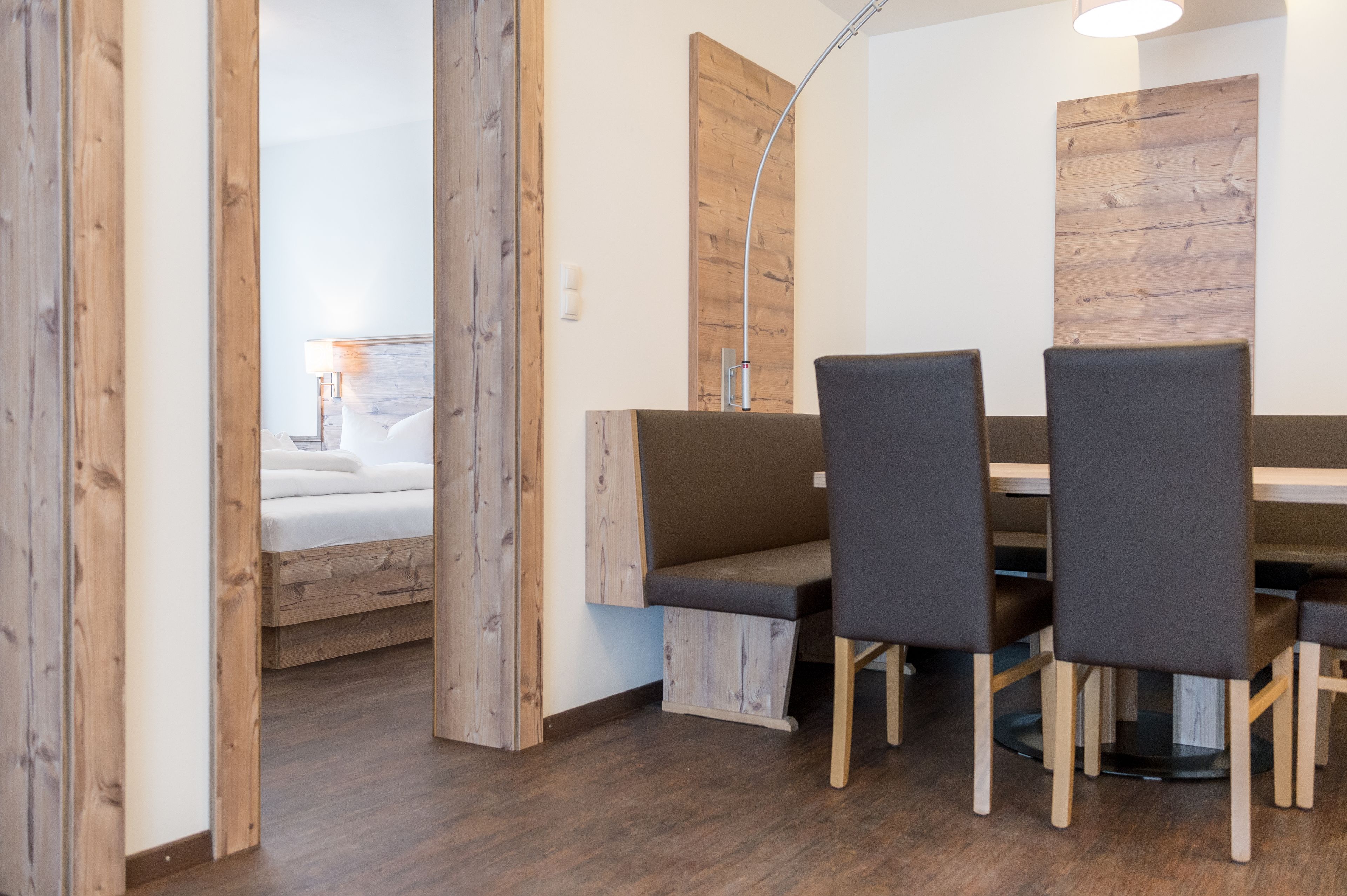 A modern room with a wooden floor, a bed with white linens, a wooden bench, and a table with four chairs.