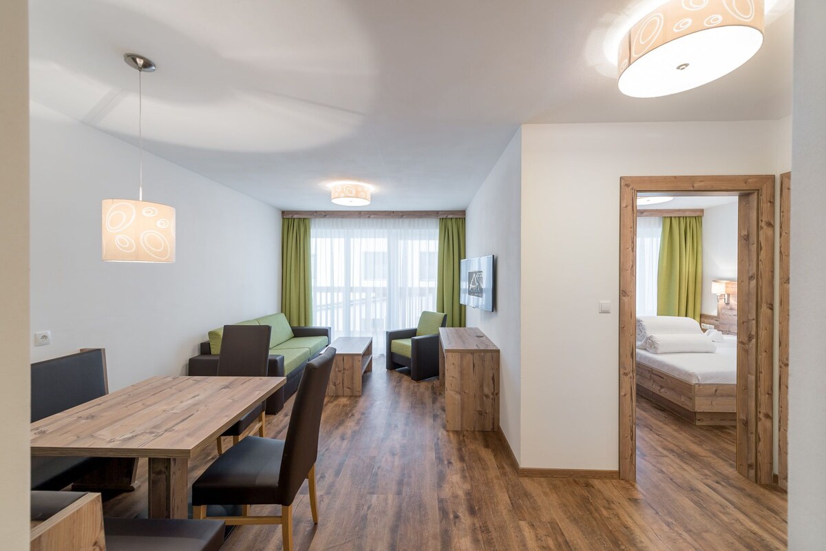 A modern hotel room with a wooden floor, a dining area with a wooden table and chairs, a sofa, a TV mounted on the wall, and a bed with a wooden headboard.