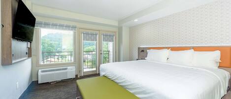 A hotel room with a large bed, a bench, a TV mounted on the wall, and a view of the outdoors through the windows.