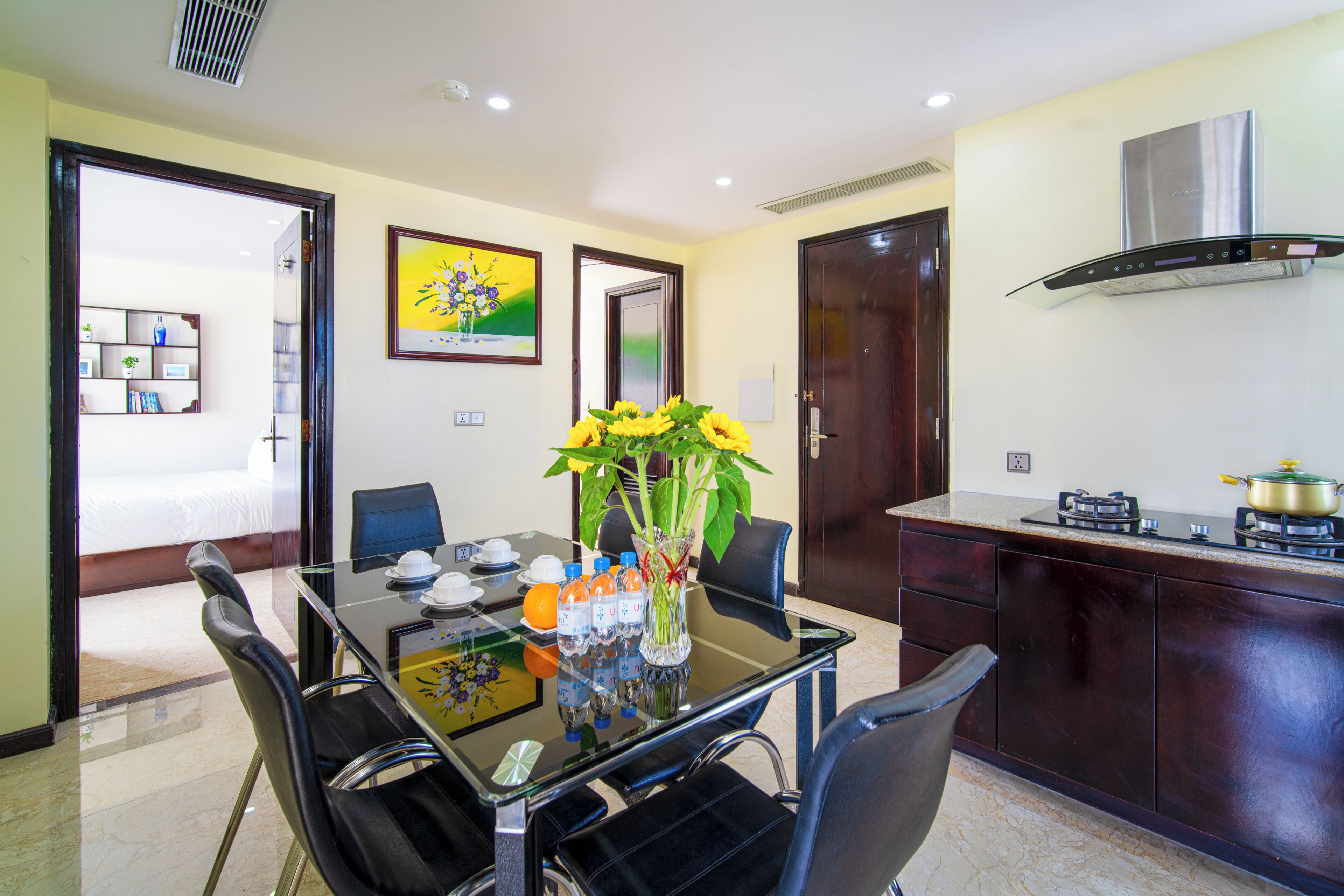 A hotel room with a dining table, chairs, a vase with flowers, a kitchen area with a stove, and a bed in the background.