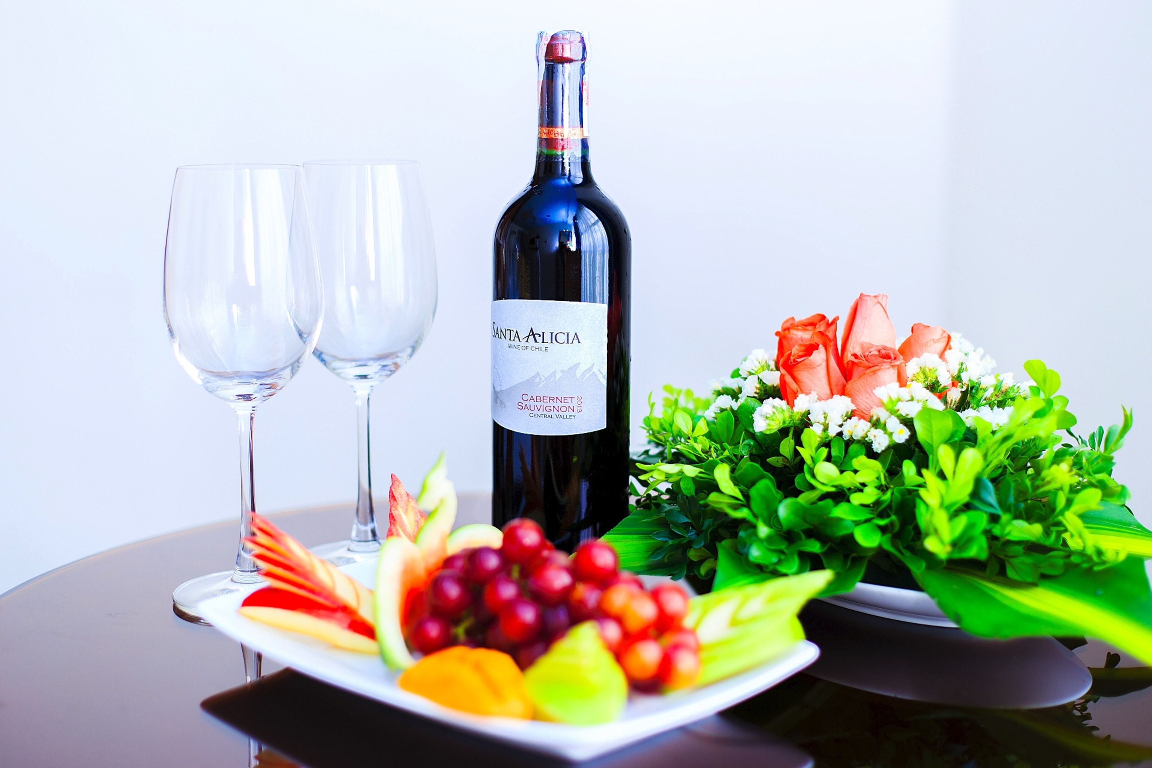 A bottle of Santa Alicia wine, two glasses, and a plate with fruit and flowers.