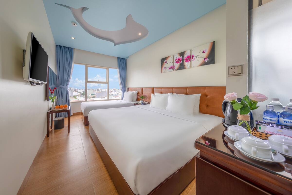 A hotel room with a large bed, a flat-screen TV, a city view, and a breakfast bar with water bottles.