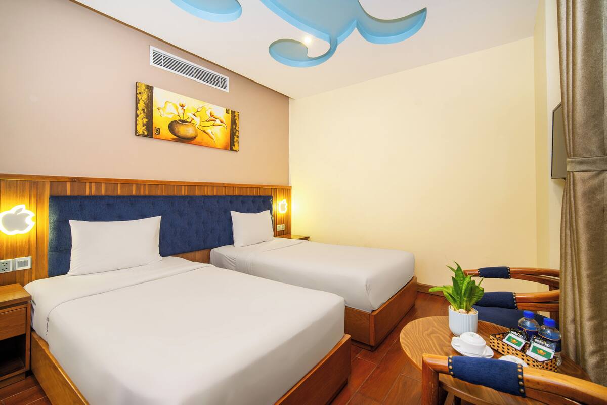 A hotel room with two beds, a wooden headboard, a wall-mounted artwork, a small table with a plant and toiletries, and a television on the right.