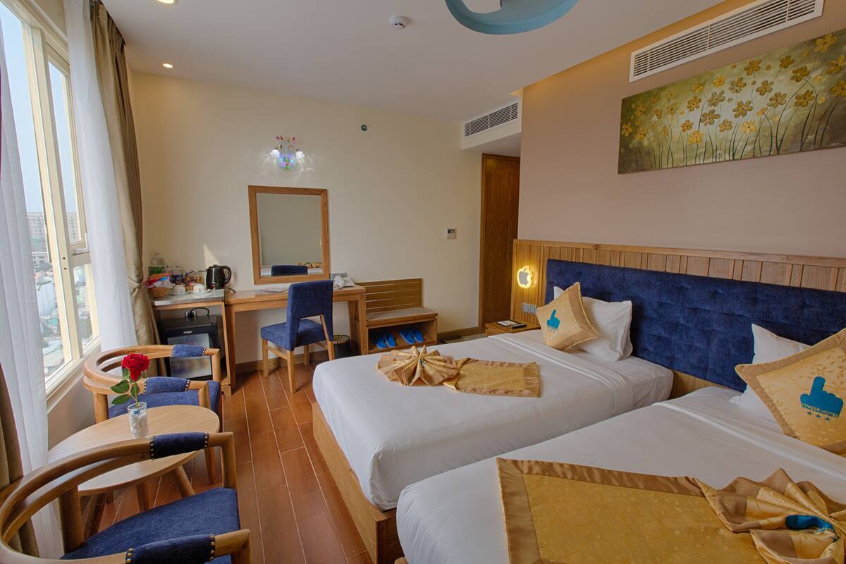 A hotel room with two beds, a desk, a chair, a small table with a vase of flowers, and a painting on the wall.