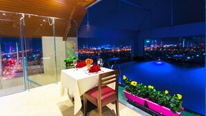 Un coin repas avec une table dressée pour deux, une vue sur la ville à travers de grandes fenêtres et une piscine visible à travers la vitre.