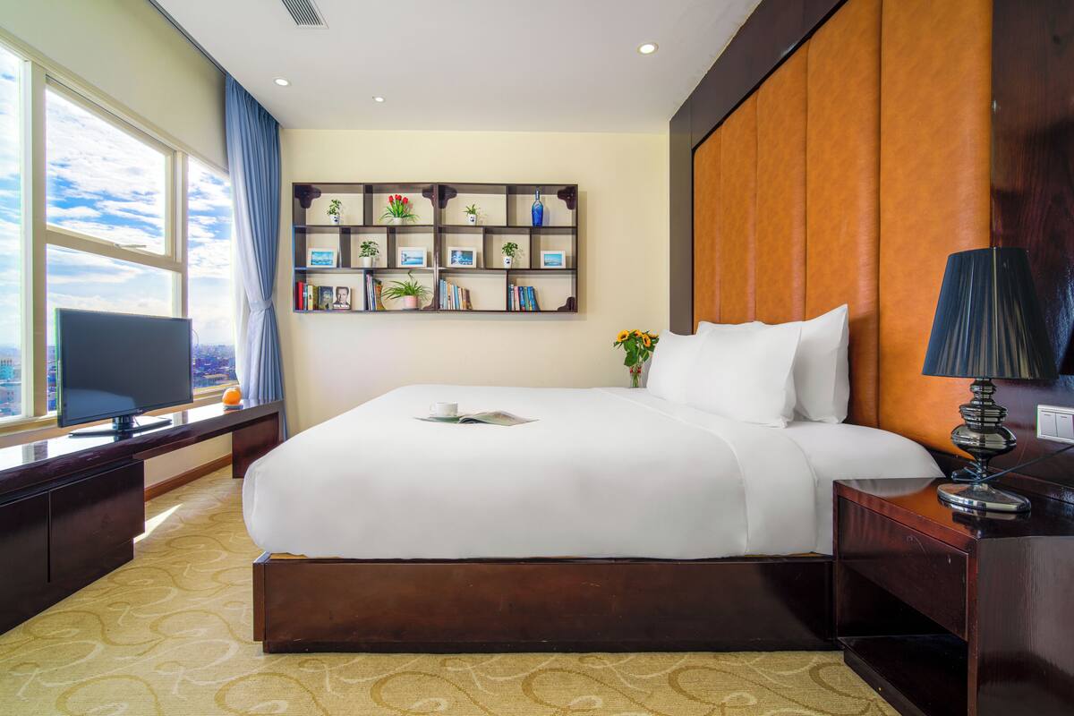 A hotel room with a large bed, a television on a desk, a bookshelf with decorative items, and a window with a city view.