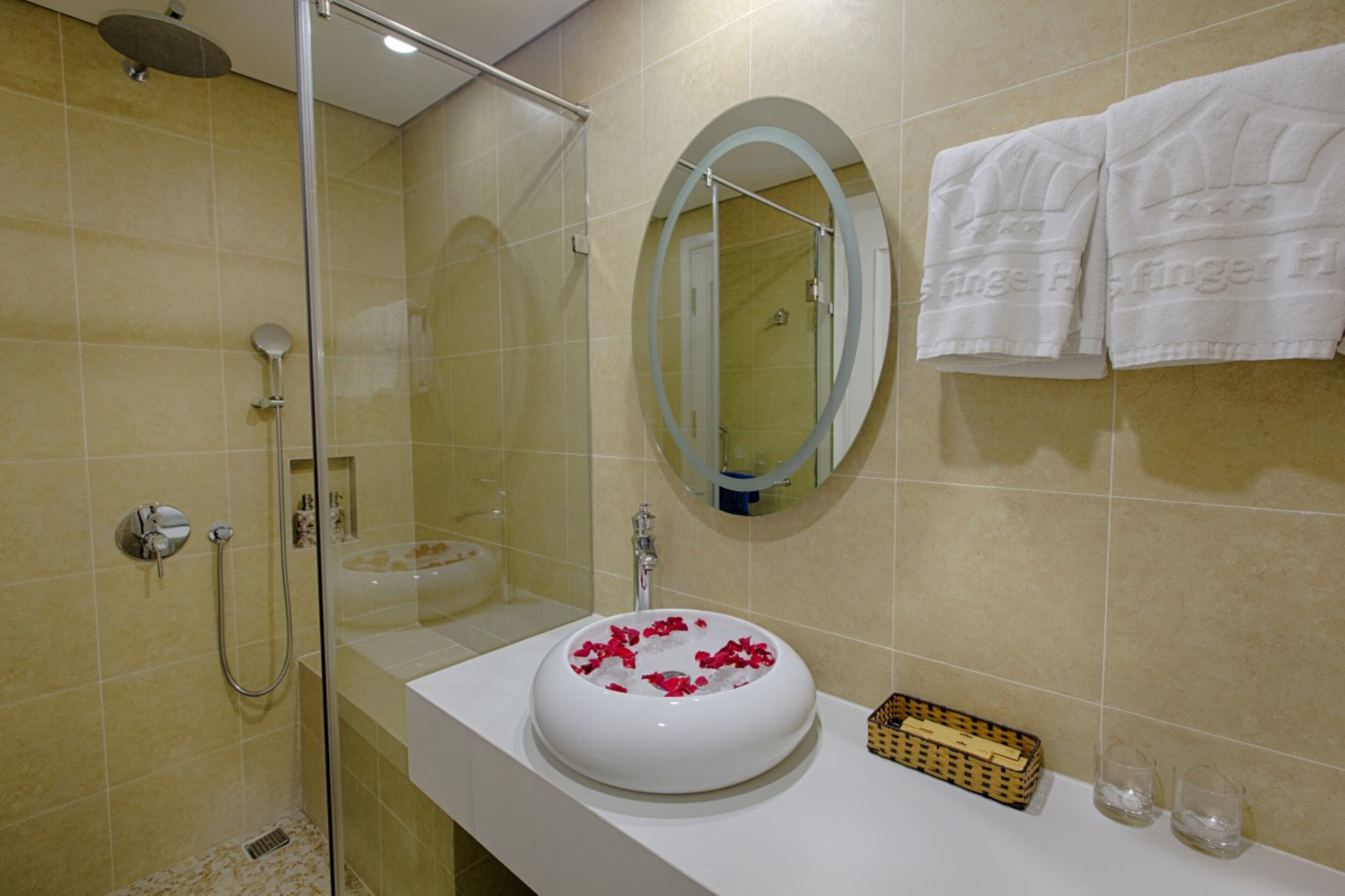 A bathroom with a glass-enclosed shower, a white sink with rose petals, and a mirror with a towel rack.