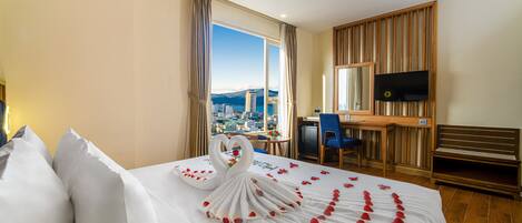 A hotel room with a bed adorned with a swan and rose petals, a desk with a chair, a television, and a view of a cityscape through large windows.