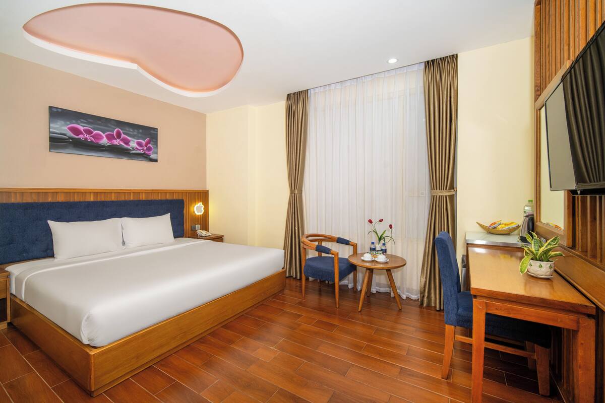 A hotel room with a bed, a desk, a chair, a small table, a potted plant, a painting, and a television.