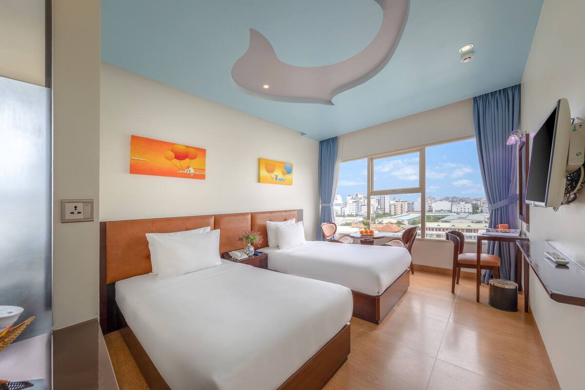 A hotel room with two beds, a desk with a chair, a television, and a large window offering a city view.