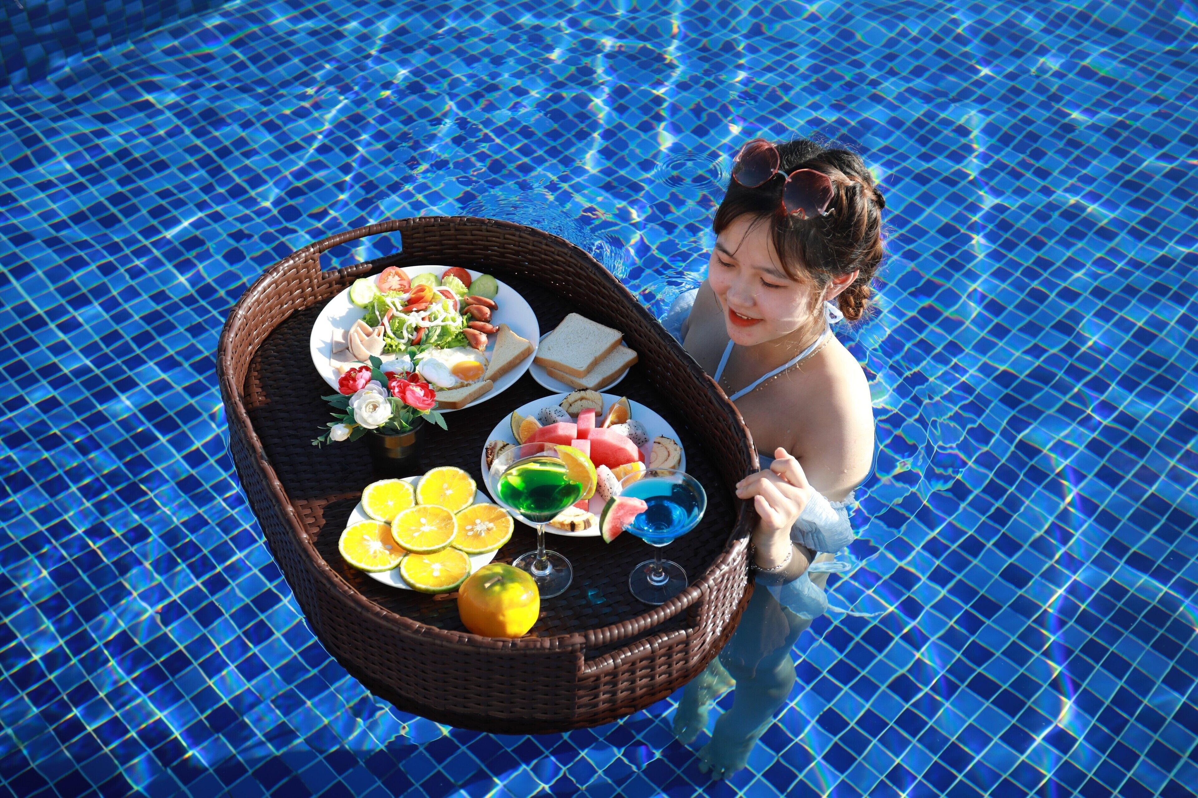 A person in a pool holding a wicker tray with food and drinks.