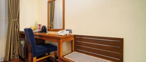 A hotel room with a wooden desk, a chair, a small table, a telephone, a mirror, a bench, and a trash bin.
