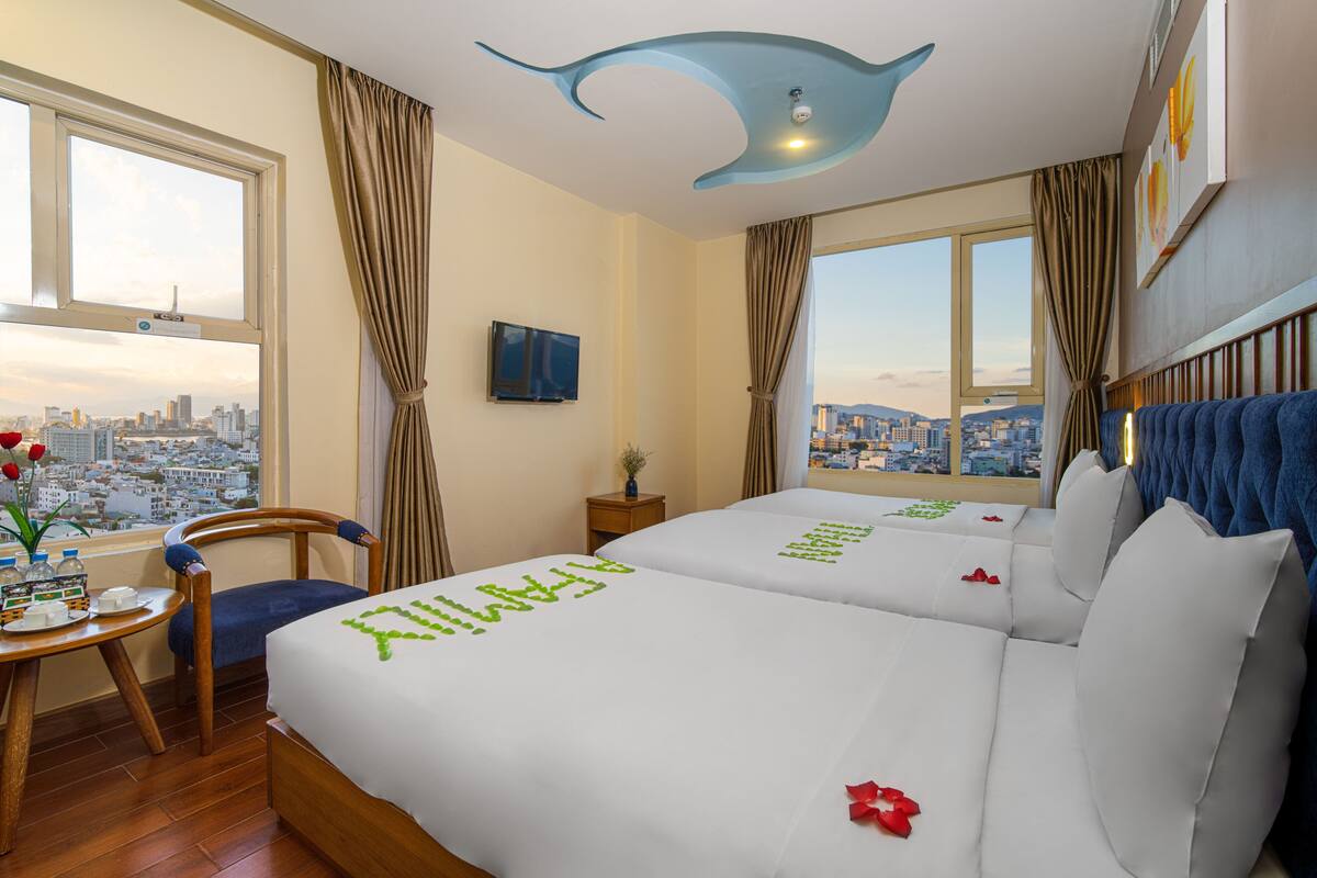 A hotel room with two beds, a city view, a desk with a chair, and a unique ceiling design.