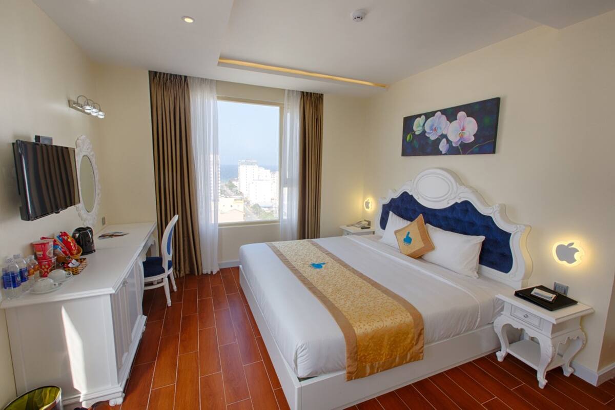 A hotel room with a bed, bedside tables, a desk with a chair, a large window with curtains, and a painting on the wall.