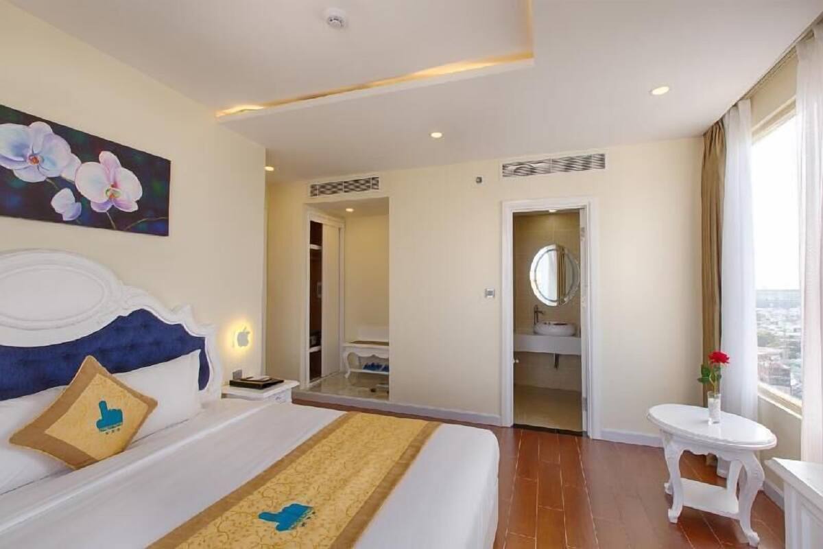 A hotel room with a bed, a bedside table, a small table with a vase, a mirror, and a view of the city.